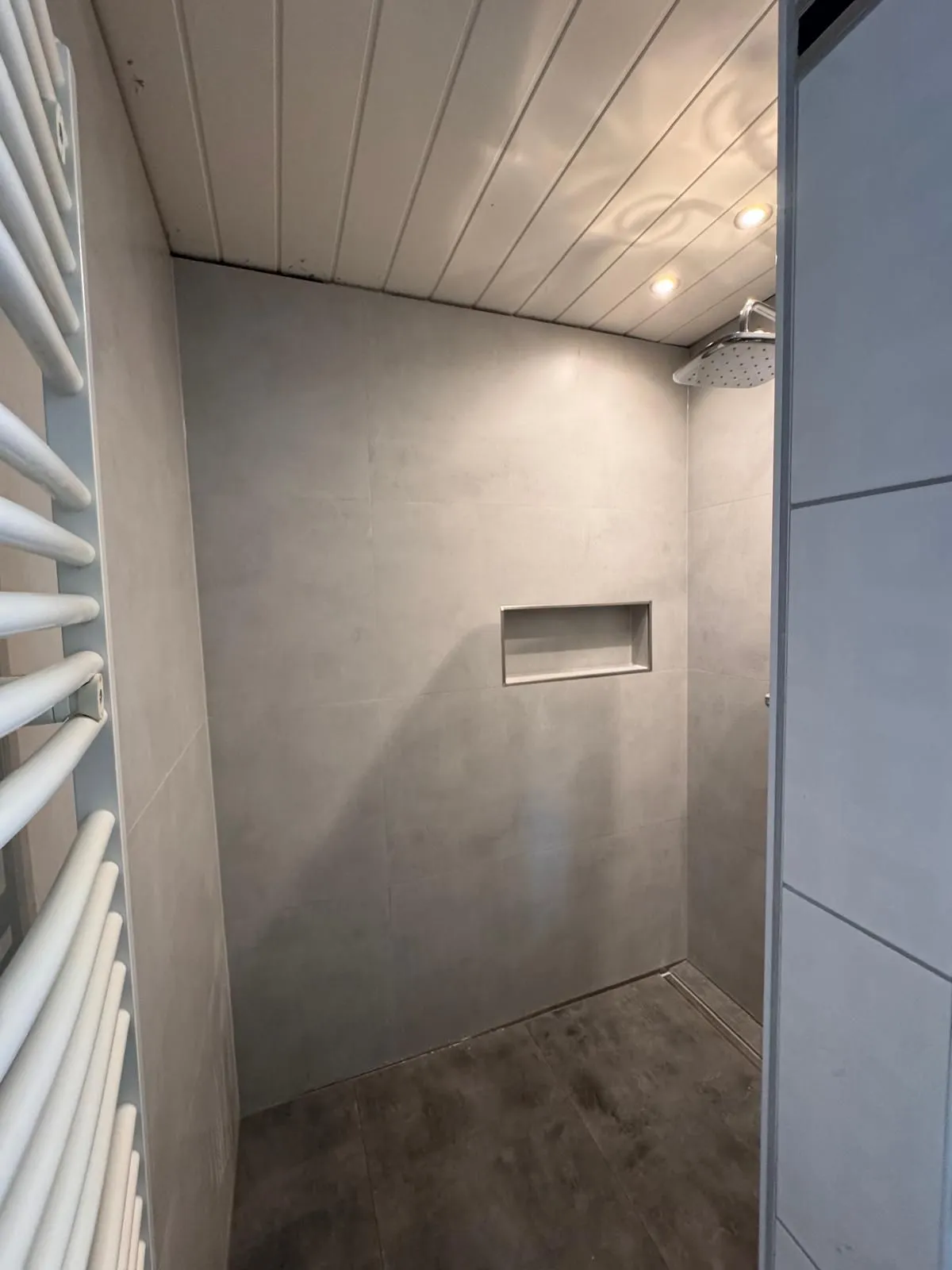 Walk-in-Dusche in Betonoptik mit eingelassener Ablagenische, Regenbrause und Duschrinne, davor ein Handtuchheizkörper.
